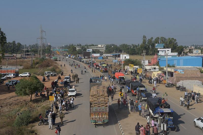 A caravan of farmers drives down a road in India. Many of the vehicles have people filling the beds of the trucks, and farmers&#039; flags hanging off the sides.