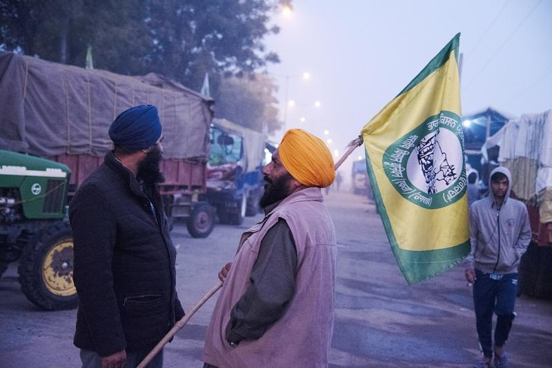 Two man stand in the road and talk at twilight. One is holding the flag of the Kisaans, or farmers, and a farm truck sits in the background.