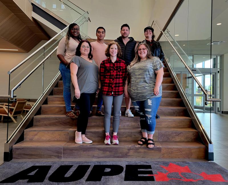 The seven members of the 2022 Young Activists Committee stand together on the main stairwell of AUPE&#039;s Edmonton Headquarters. A mat at the bottom of the stairs has an AUPE logo.