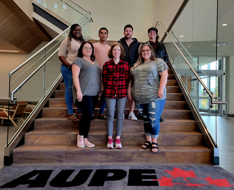 The seven members of the 2022 Young Activists Committee stand together on the main stairwell of AUPE's Edmonton Headquarters. A mat at the bottom of the stairs has an AUPE logo.